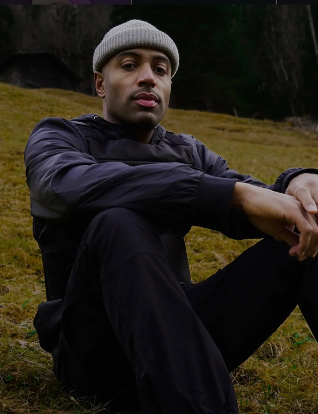 DJ Platinum sitting on a grassy slope in a beanie, forest behind.