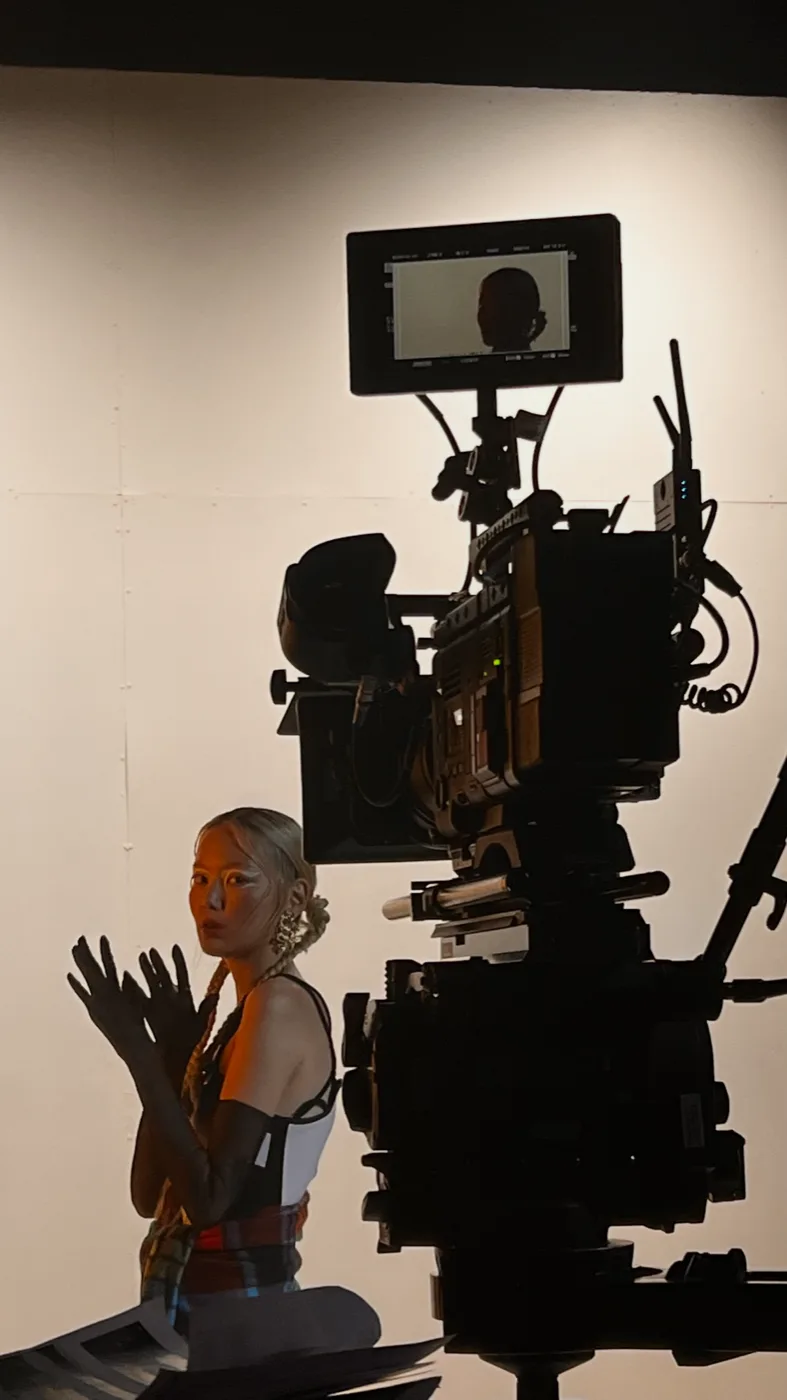 A cinema camera on set, monitor above it, a performer waiting in the light behind.
