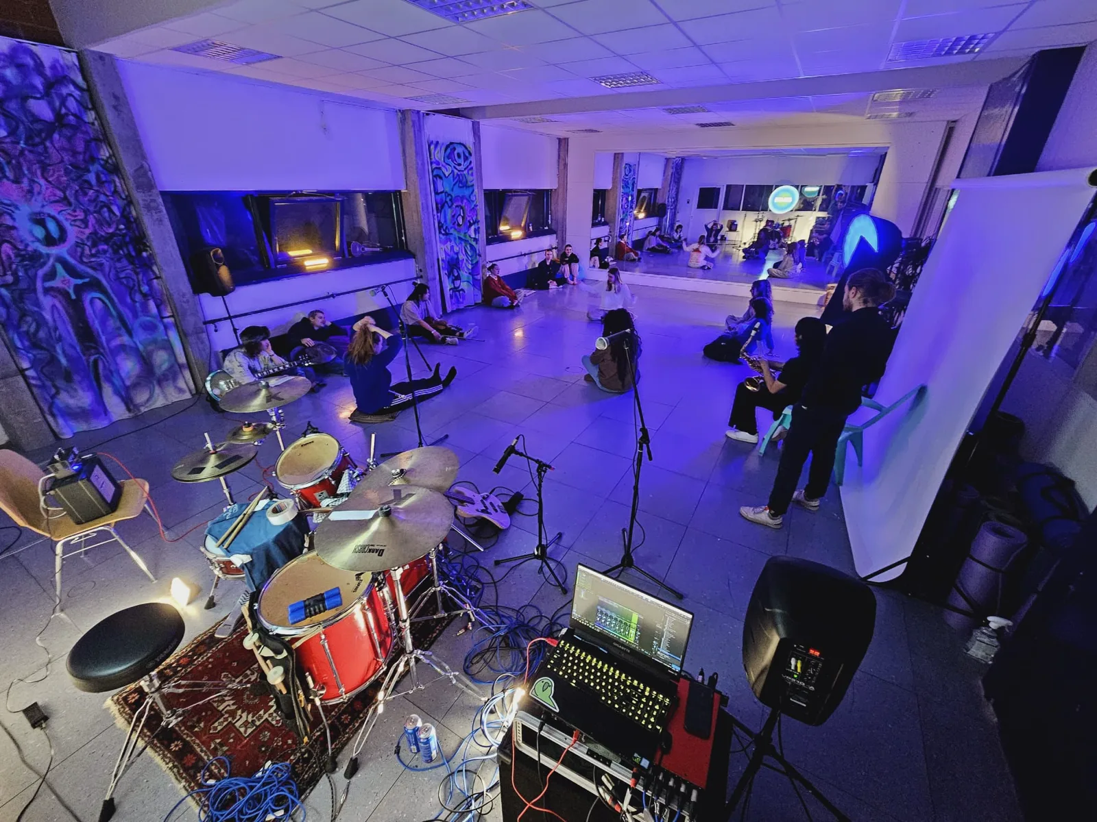 A jam session in the studio under blue light, drums and guitar set up, people sitting along the mirrors.