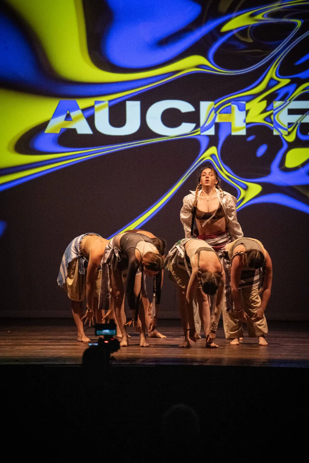 Dancers folded forward in a line, one standing, the AUCH logo projected behind them.